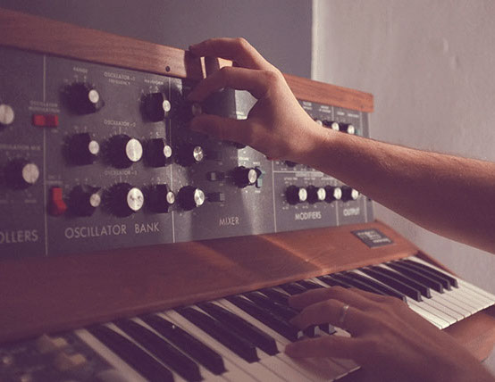 Photo couleur d'un personne qui joue sur un synthétiseur et qui manipule des boutons. Détail des mains et de l'instrument.